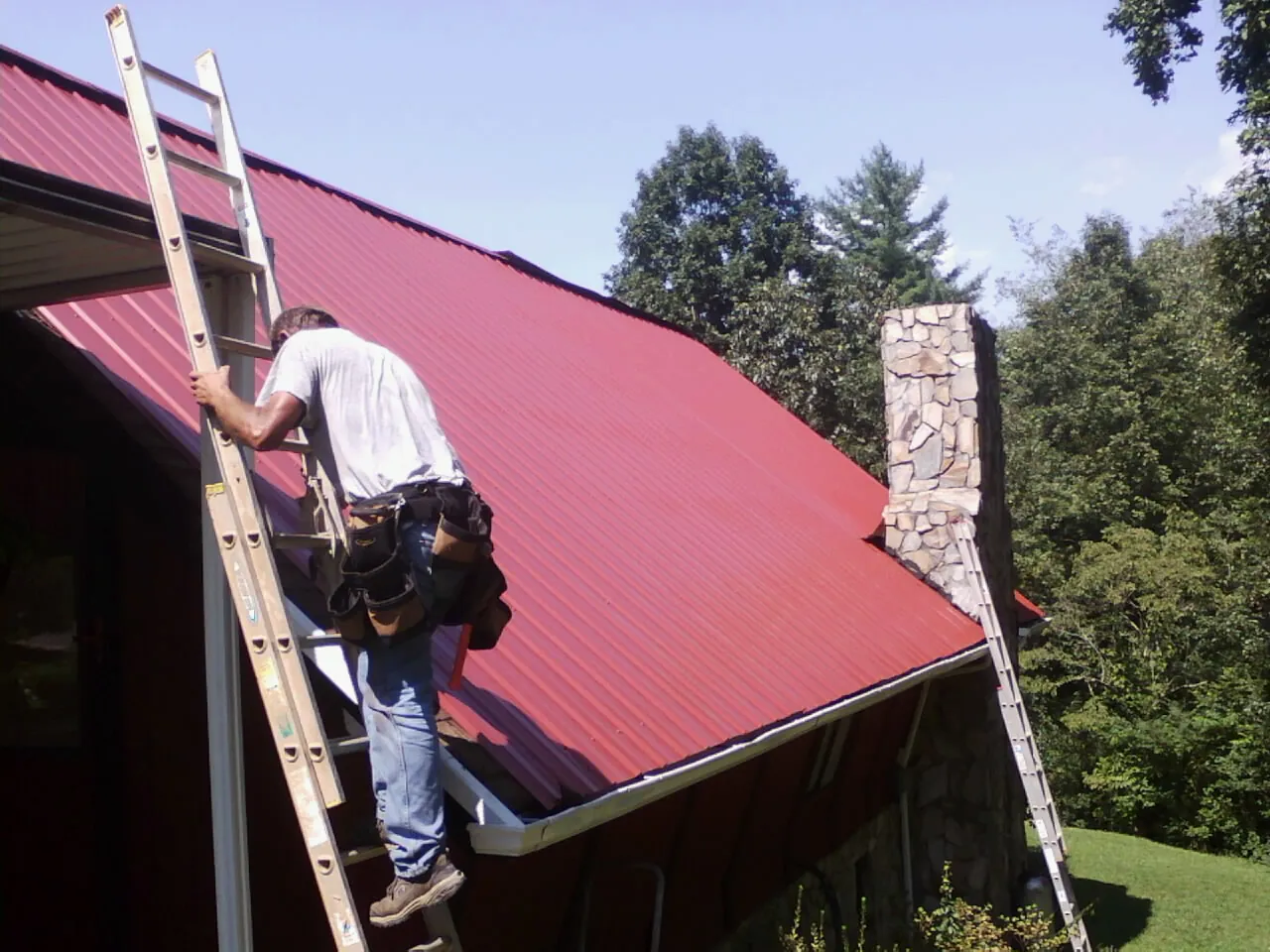 Roofer on a ladder beside a red metal roof and stone chimney — Soffit & Fascia Repair by Roofing Brothers in Fremont